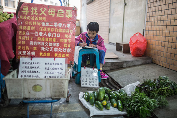 Mao Mao does her homework as she sells vegetables on a street in Dongguan, Guangdong province, on Friday. A poster beside her reads: Dear Momand Dad... I am looking forward to seeing you and having you take me home.(Zhang Youqiang/For China Daily)