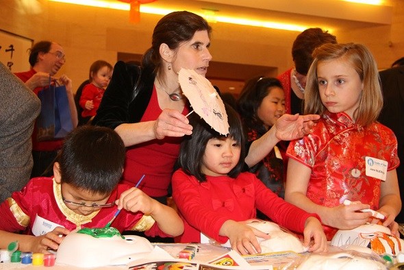 Miriam Mintzer from Maryland holds a paper umbrella while her adopted son, Eli, (seated, left) and other children paint masks at the Chinese Embassy in Washington, on Feb 12, 2018. A Chinese New Year reception was held there for American families who have adopted Chinese children. The event was supported by the Cradle of Hope Adoption Center and the Barker Adoption Foundation in the United States. (Zhao Huanxin / China Daily)