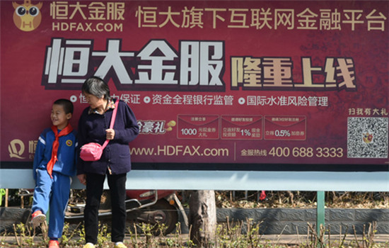 A grandmother and her grandson wait for a bus in front of a billboard of Evergrande Financial advertising, its new online services, in Kunming, capital of Yunnan province.(Photo by Li Ming/For China Daily)