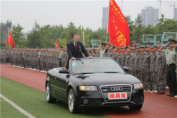 The university president Shi Xiuhe stands in a black open-roof Audi complete with microphone inspecting students wearing olive-green camouflage uniforms, alongside saluting military instructors.