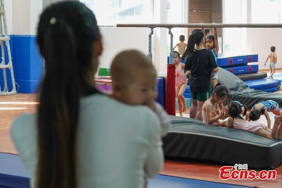 A mother holds her child as she watches young gymnasts train at the Amateur Children\'s Gymnastics Training School in Rongjiang County, Southwest China’s Guizhou Province, July 16, 2018. There are 60 children taking part in gymnastics training at the school during their spare time. Since 1972, the county has sent 21 athletes to the provincial gymnastics and gymnastics trampoline teams, among whom six later became national team members. The county is home to Liu Rongbing, who won a gold medal for the men\'s team in the 2014 World Artistic Gymnastics Championships. (Photo: China News Service/He Junyi)