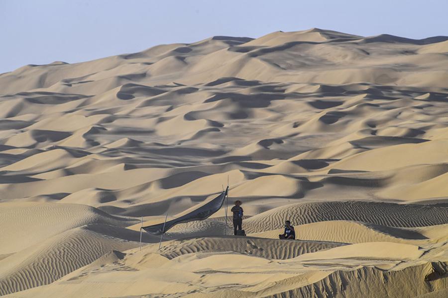 On a sand dune, two workers have a break near a temporary mat shelter.  (Photo/Xinhua) 
Workers are building only the third paved artery through the Taklimakan Desert to help people and products move more freely

Five bulldozers were working on the side of a 9-meter-high dune. In one of them sat Zhang Xiaodong, his cab swirling with sand. Fine dust had collected in the lines of his sweaty face.

The construction team is building a new road through the Taklimakan Desert, the largest desert in China, covering 337,000 square kilometers in the Xinjiang Uygur autonomous region.

When it is finished, the road will connect the counties of Qiemo in the north and Yuli in the south, becoming only the third paved artery through the vast expanse known as the \