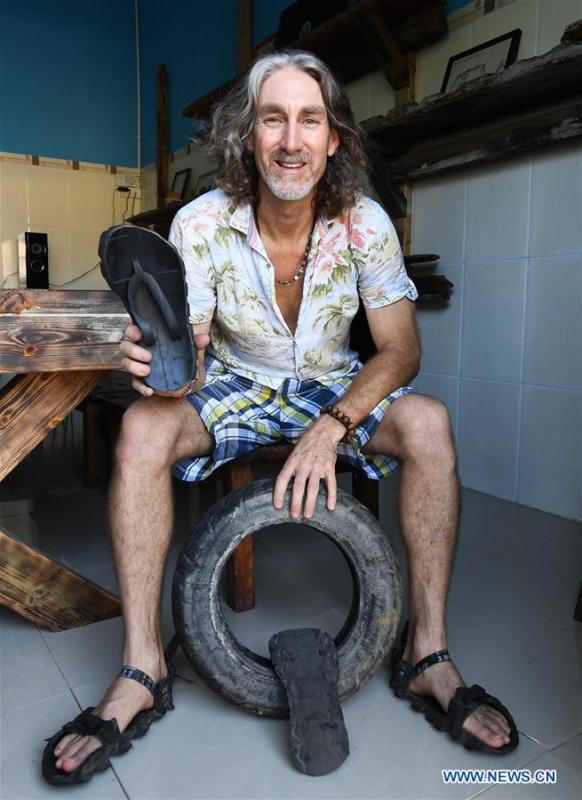 Craig Anderson shows slippers made from an old tire at the eco-sustainability service center of the West Island in Sanya, south China\'s Hainan Province, Sept. 19, 2018. Anderson arrived in February at the West Island inhabited by around 4,000 people. Locals regarded Anderson as a scavenger when he first arrived, as he wandered around the beach almost every day, picking up driftwood, plastic bottles, shattered glasses, and rusty pieces of iron. As time went by, the locals realized that Anderson is an artist who devotes himself in environment protection art design. He profits the recyclable and regenerative materials in making art works. (Xinhua/Yang Guanyu)